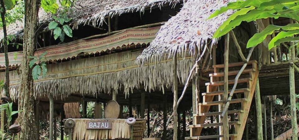 A couple standing in front of a traditional wooden house on stilts with a thatched roof and a sign that says “Rumah Murut” at Mari-Mari Cultural Village, a cultural attraction in Sabah that showcases the history and lifestyle of five ethnic tribes.