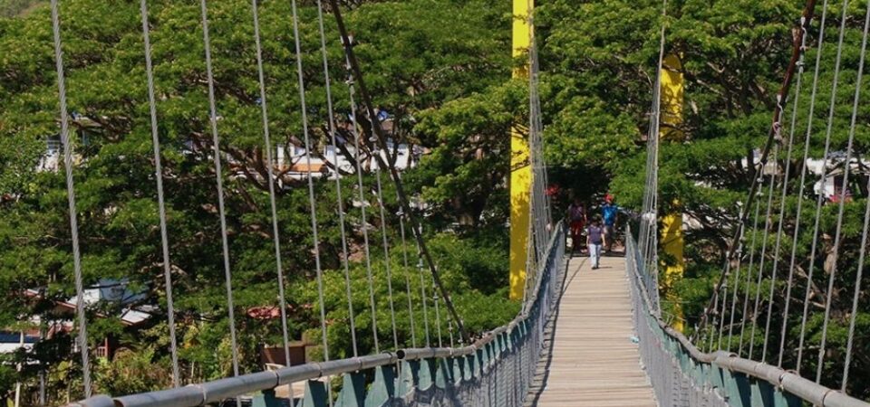 A couple walking on a concrete bridge over a yellow river with a sign that says “Jambatan Tamparuli” at the entrance of the bridge, a famous landmark in Sabah that has a tragic legend behind its construction.