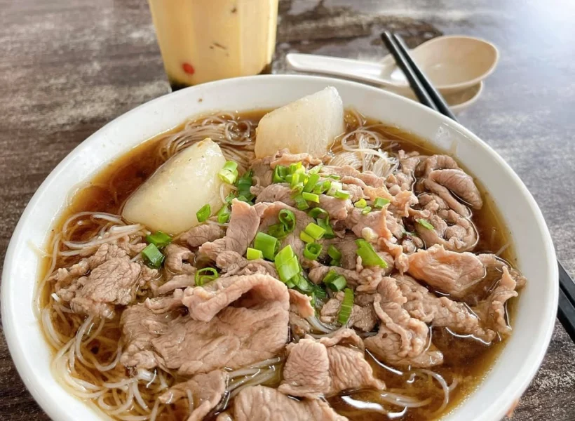Bowl of Ngiu Chap in Sandakan, filled with noodles, beef slices, meatballs, and herbs in a rich brown broth.